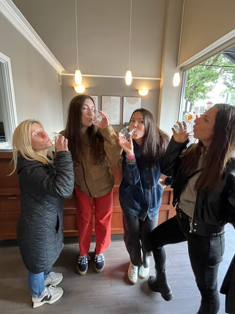 ladies drinking at Windemere Four women stand indoors near a counter, each holding and drinking from a small glass, with hanging lights and a window in the background.