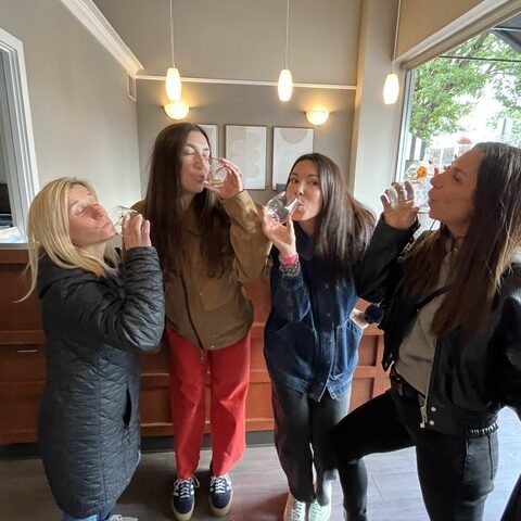 Four women stand indoors near a counter, each holding and drinking from a small glass, with hanging lights and a window in the background.